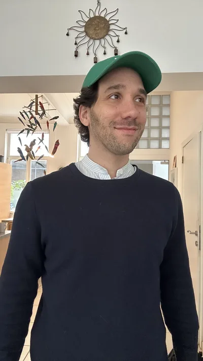 Ma photo de départ : debout chez moi, pull marine sur une chemise rayée, casquette verte